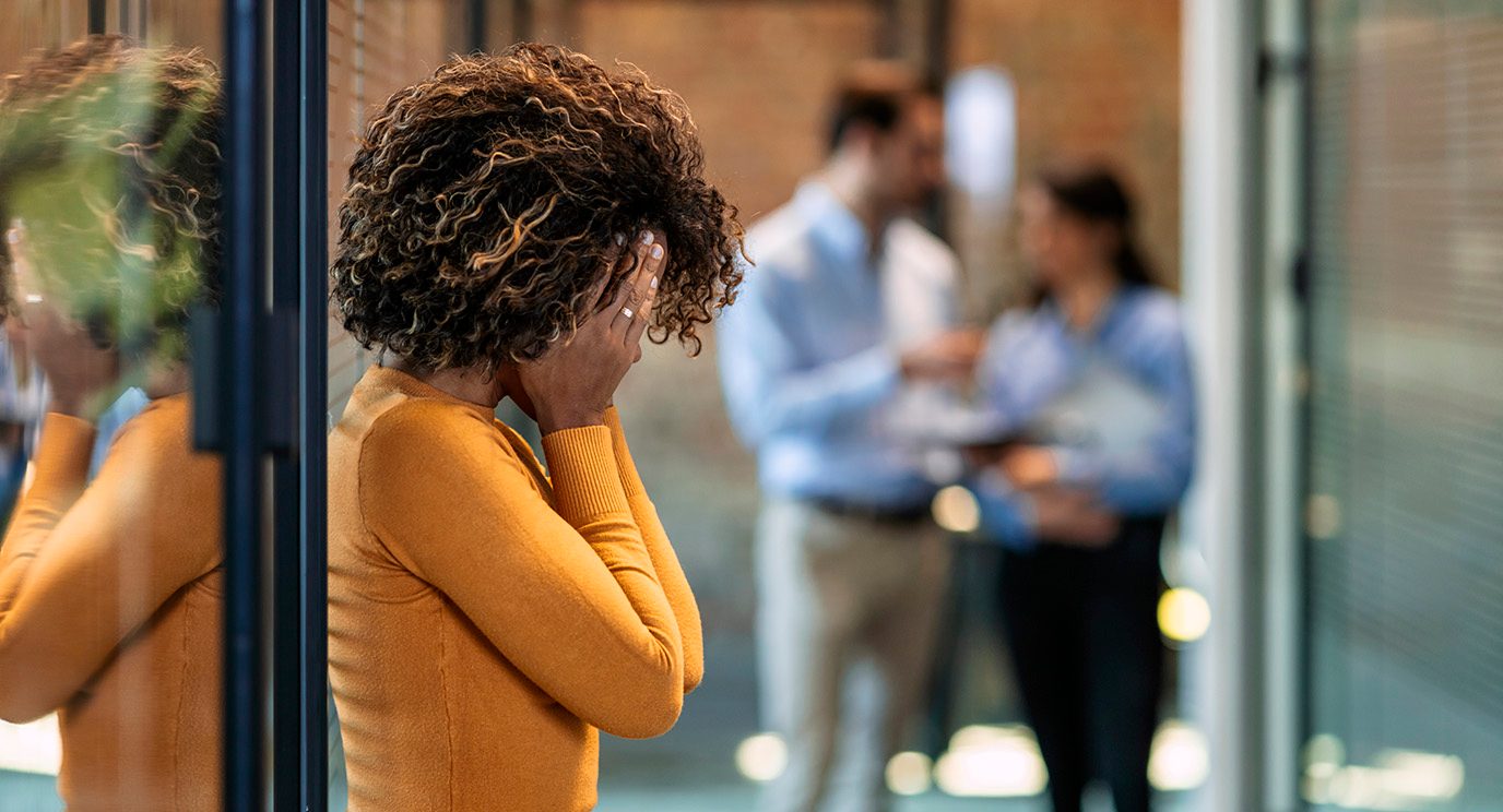 Black woman in orange blouse covering face in distress in an office hallway
