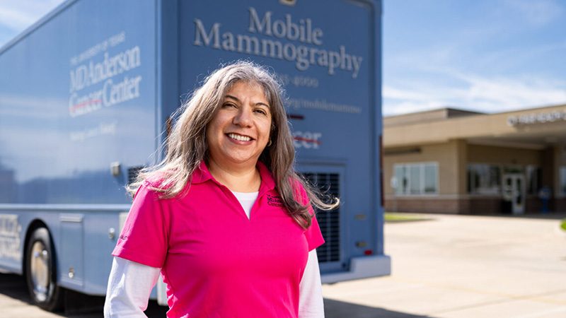 Lucy Balderas, a woman with long brown hair wearing a pink polo shirt with a white long sleeve underneath, smiles in front of a blue mobile mammography van