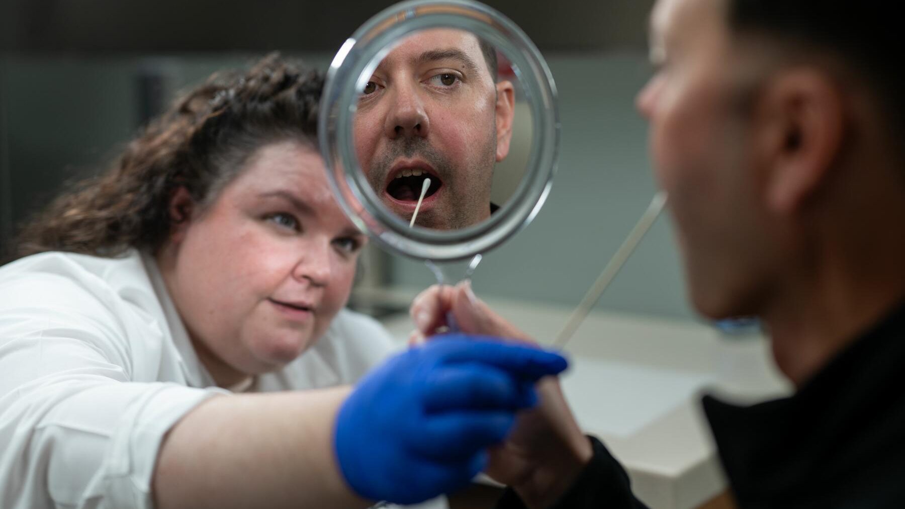 A clinician wearing a blue medical glove holds a cotton swab near the mouth of a man holding a mirror.