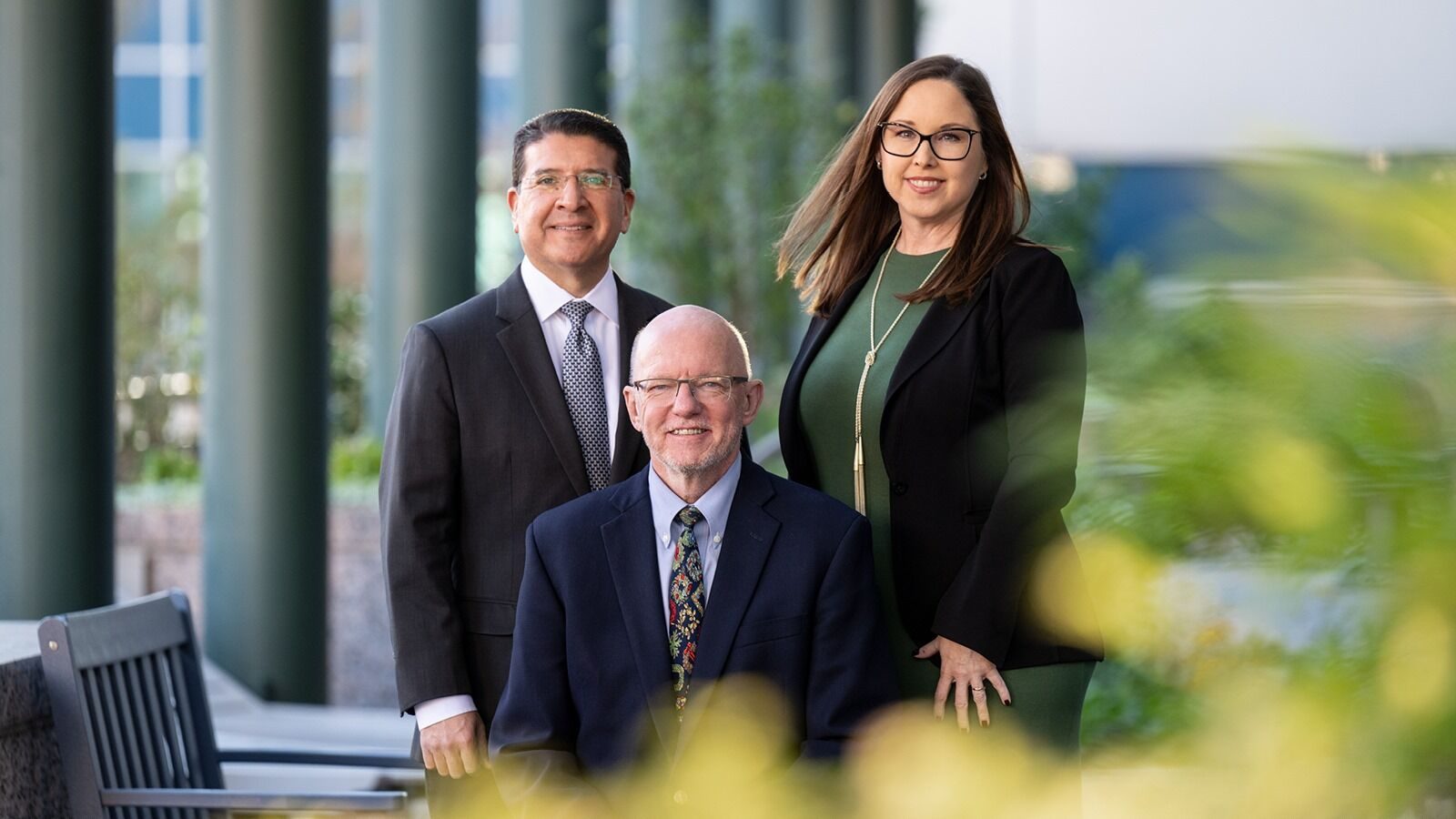 A man in a suit sit in front of a standing woman in a green dress and blazer and a man in a black suit.