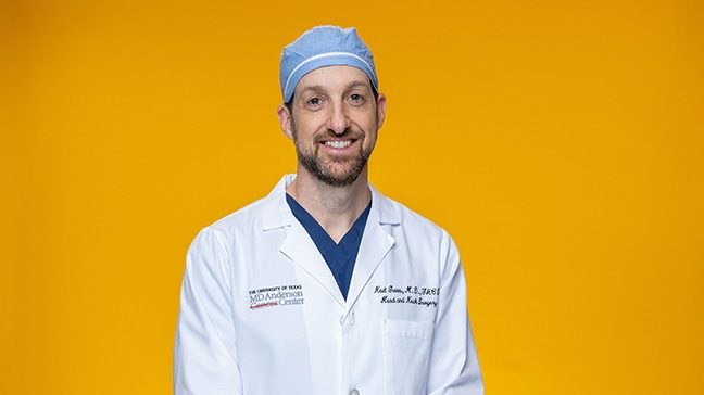 Man in white coat, navy scrubs and light blue surgical cap with a bright orange backdrop.