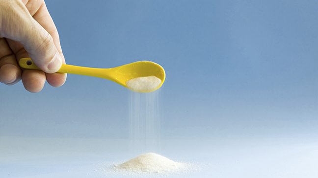 Hand holds a yellow spoon pouring out white granulated sugar