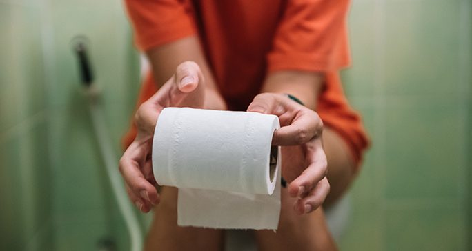 Female in an orange shirt sitting on the toilet holding a roll of toilet paper