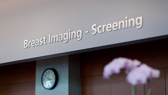 A grey wall with an analog clock and a sign below a sign reading 'Breast Imaging - Screening'