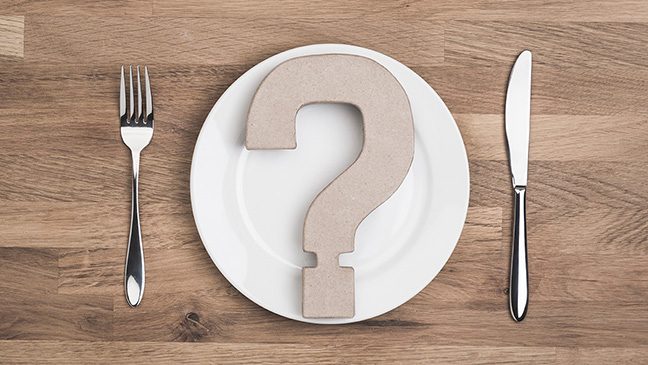 A white plate with a cardboard question mark placed on it sits on a wooden surface. A silver fork and knife sit on either side of the plate.