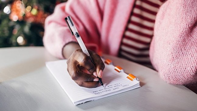A person wearing a pink sweater and red striped shirt holds a pen and writes in a notebook