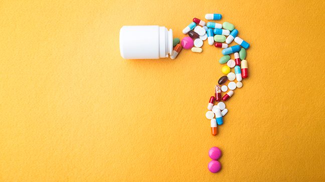 White pill bottle spills out a variety of colorful capsules and pills in the shape of a question mark against an orange background