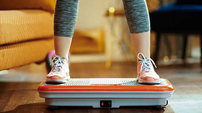 Person wearing gray leggings and orange tennis shoes stands on an orange and gray vibration plate placed in front of an armchair.