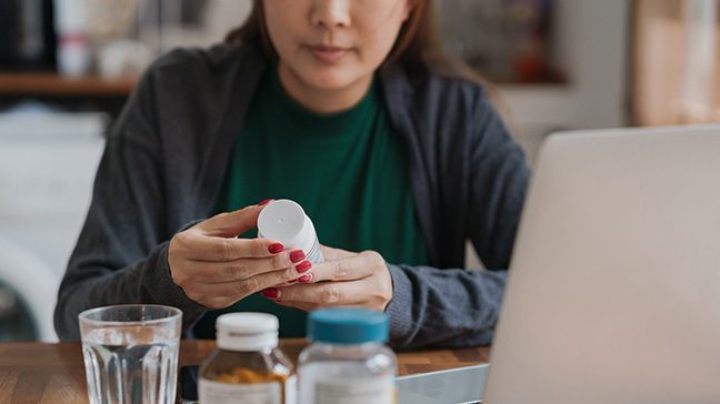 Woman holds white pill bottle. On the table in front of her there is a laptop, glass of water and two additional pill bottles.