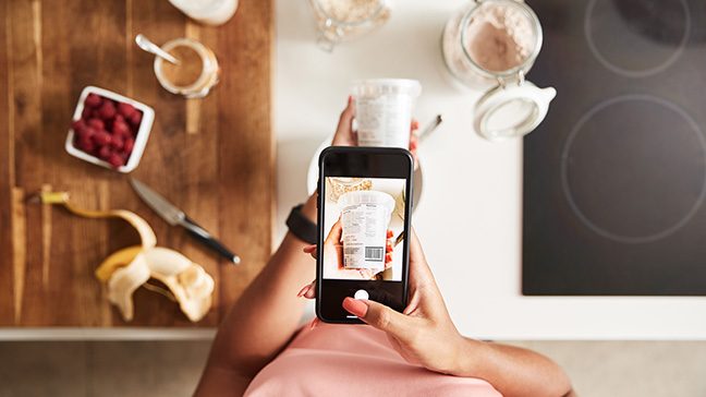 Overhead view of person using smart phone to take picture of the nutrition label of a yogurt container while preparing breakfast in the kitchen.