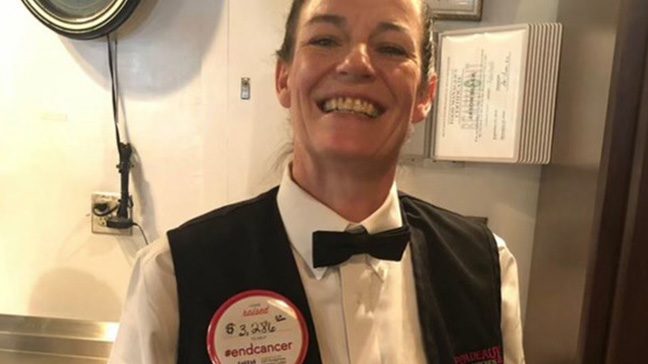 Kelli is shown smiling at work at Pappas restaurant wearing an end cancer sticker as part of the Pappas MD Anderson fundraising campaign