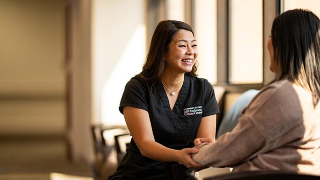 A woman in black scrubs smiles and holds the hand of the woman she is speaking with.