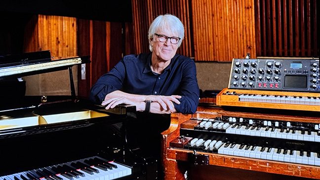 Man wearing glasses and button down shirt poses between a piano and a keyboard.