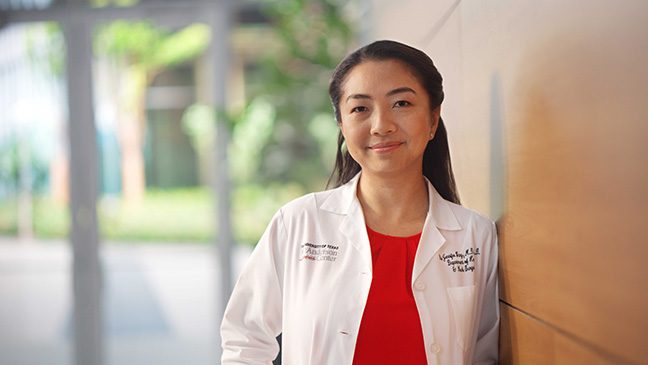 Brunette woman in red blouse and white coat stands in a hallway with a window behind her.