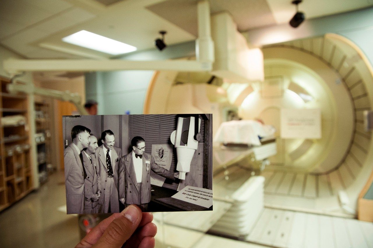 Dated photo of MD Anderson doctors looking at a model of an irradiator juxtaposed in front of a current proton therapy machine.