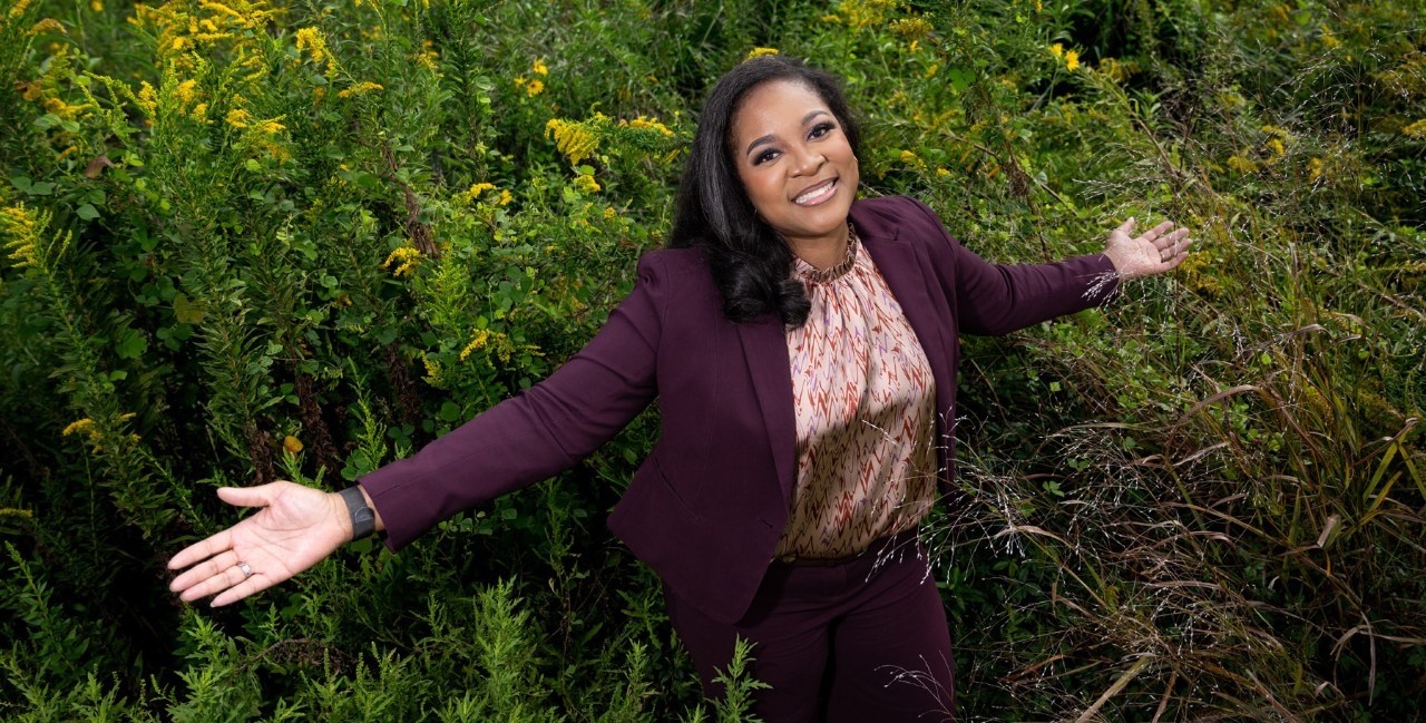 A smiling woman in a purple cardigan stands with her arms outstretched in a field of green plants