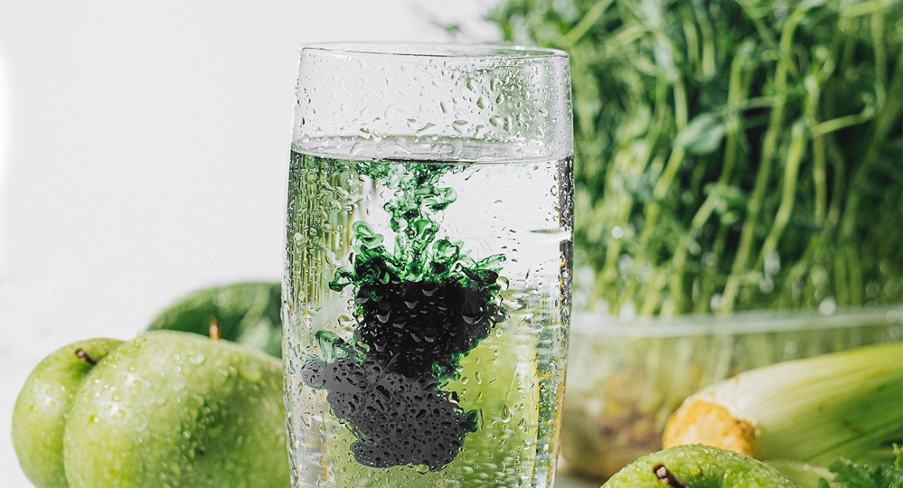 Glass of water with chlorophyll drops in it. Green fruits and vegetables are shown behind.