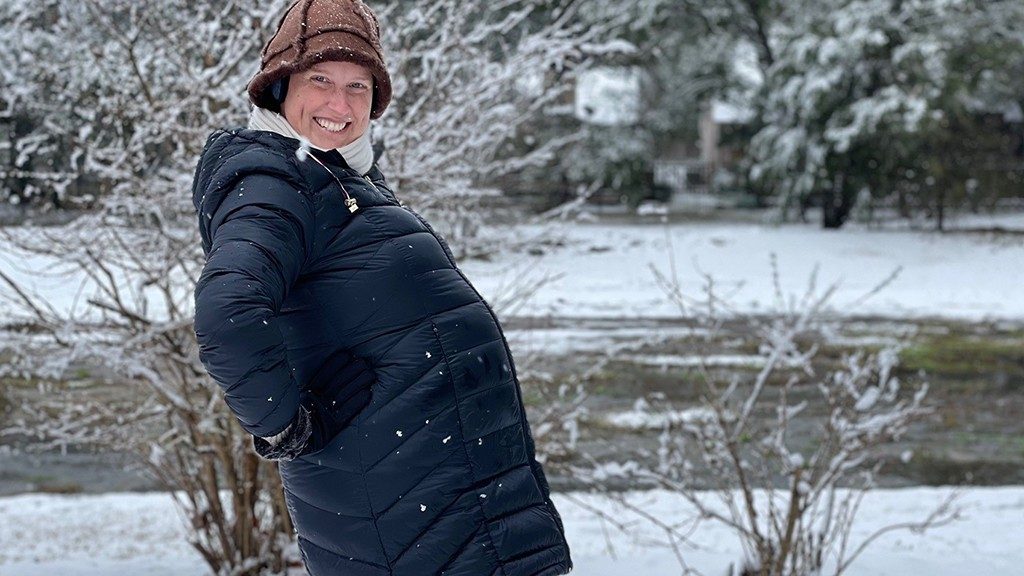 Photo of triple-negative inflammatory breast cancer survivor Alessandra Land wearing her winter coat and hat in the snow while showing her pregnant belly