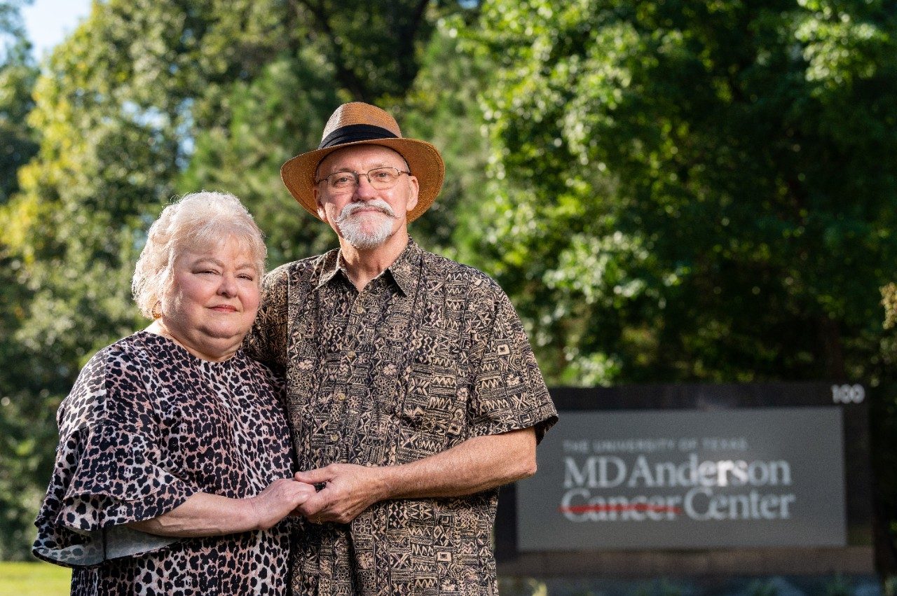 Breast cancer survivors Teresa and Billy Mayo stand holding hands beside and MD Anderson Cancer Center sign.