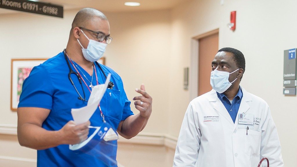 Josiah Halm, M.D. (right, wearing a white coat and face mask) chats with Advanced Practice Nurse Andre Brantley (wearing scrubs and a medical-grade face mask) as they walk down the hall in the clinic, wearing masks