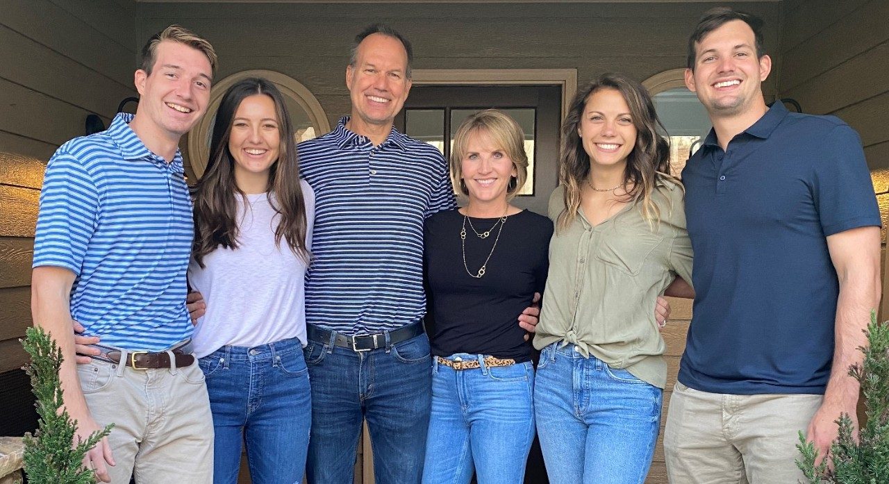 Elizabeth Somers stands in the middle in a photo with two daughters, two sons and her husband to her left. All are wearing pants and a cotton shirt and smiling outside  in front of a house.