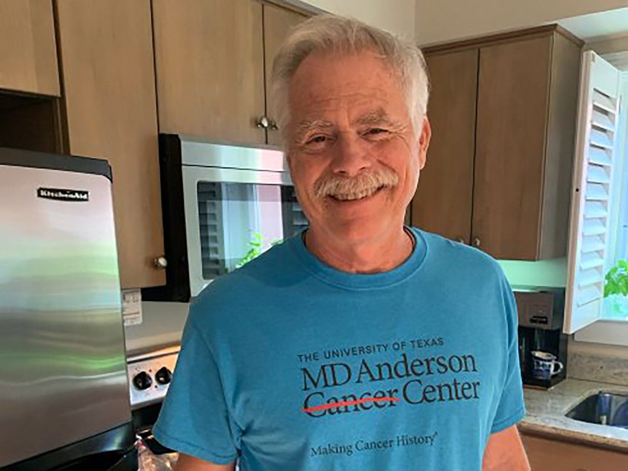 Patrick Mead smiles in his kitchen while wearing a blue t-shirt with the MD Anderson logo on it. He is an older white man.