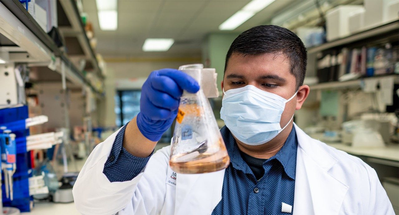 Graduate student Rolando Vedia holds a  beaker in the lab while wearing a white coat, medical-grade face mask and blue gloves.