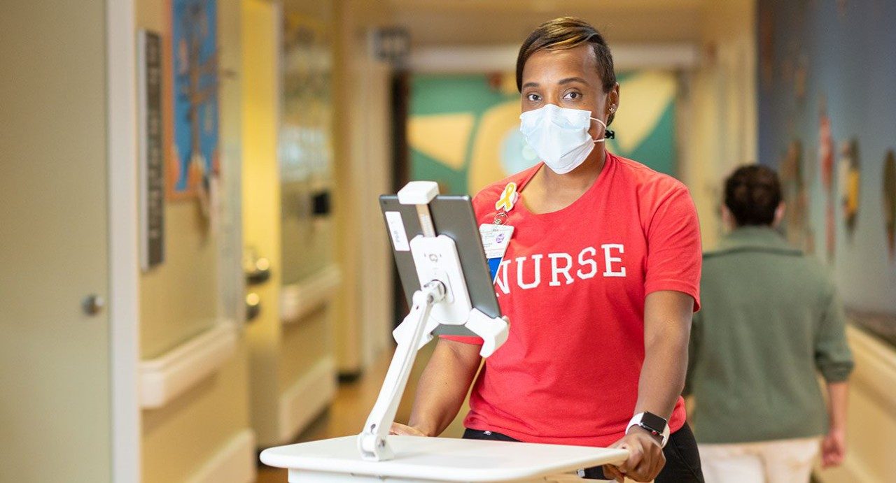 Hospital nurse Tiambe Kuykendall stands in front of an iPad in the hallway at MD Anderson while wearing a medical-grade face mask and red t-shirt that says "Nurse." 