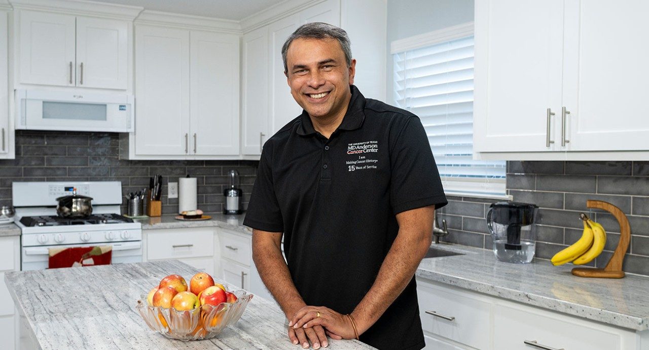 Ronald Abraham stands in his newly renovated kitchen. He has his hands on a white island, while a bowl of red apples sits beside him. He is smiling and wearing a black collared shirt with the MD Anderson logo in the upper right corner.