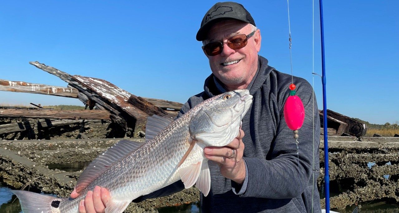 Bob Seibert holding up a fish he caught, while a lake and greenery are behind him. He is wearing sunglasses and a hat. 