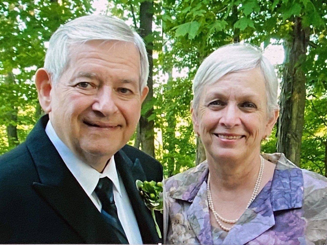 Bob Wagar, M.D.,  and his wife, Robin stand in front of green trees while wearing a suit and a purple and pink floral blazer and pearls, respectively. They are an older white couple with gray hair.
