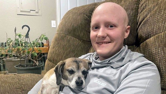 Connor Johnson relaxing in easy chair with his dog, Emma. Connor's head is bald, and he is smiling.