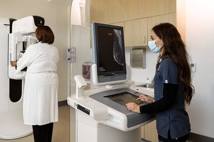 Person standing in front of a diagnostic imaging screen viewing results of another person in the room receiving a mammogram