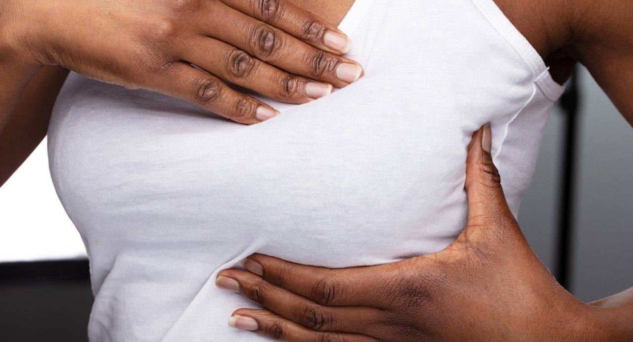 Close-up view of two Black hands palpating their owner's breast beneath a white tank top