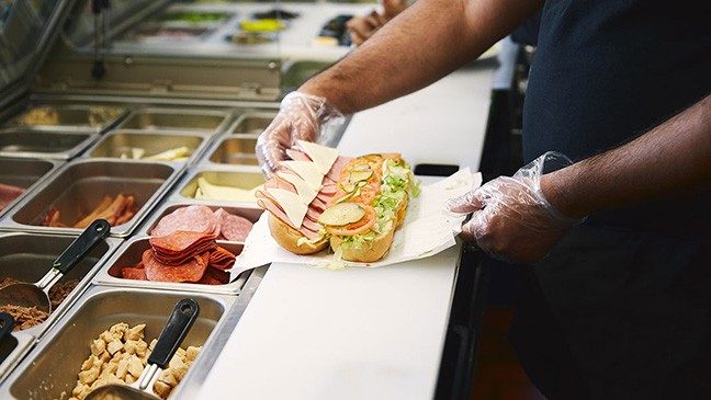 A person wearing plastic gloves assembles a sandwich with ham, cheese, tomatoes, pickles and lettuce.
