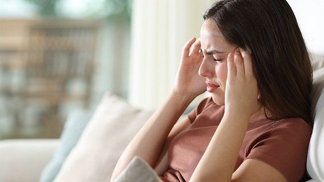 A grimacing woman sits on a couch with her hands on her temples.