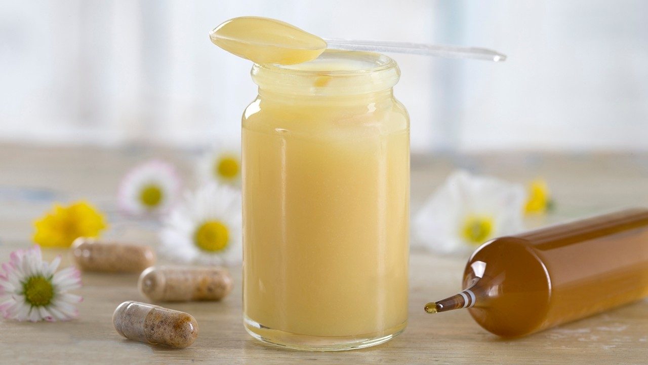 A jar of yellow royal jelly with a spoon resting on top sits on a counter alongside three supplement capsules, white flowers and a tube of royal jelly.