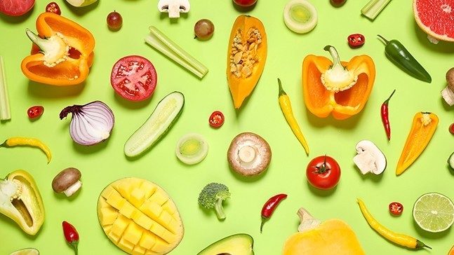 Whole and sliced fruits and vegetables on a lime green background including onion, grapes, broccoli, cucumber, tomatoes, mushroom, squash, orange peppers, chili peppers, limes, mango, zucchini and grapefruit.