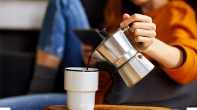 A person in an orange shirt pours coffee from a silver moka pot into a white mug.