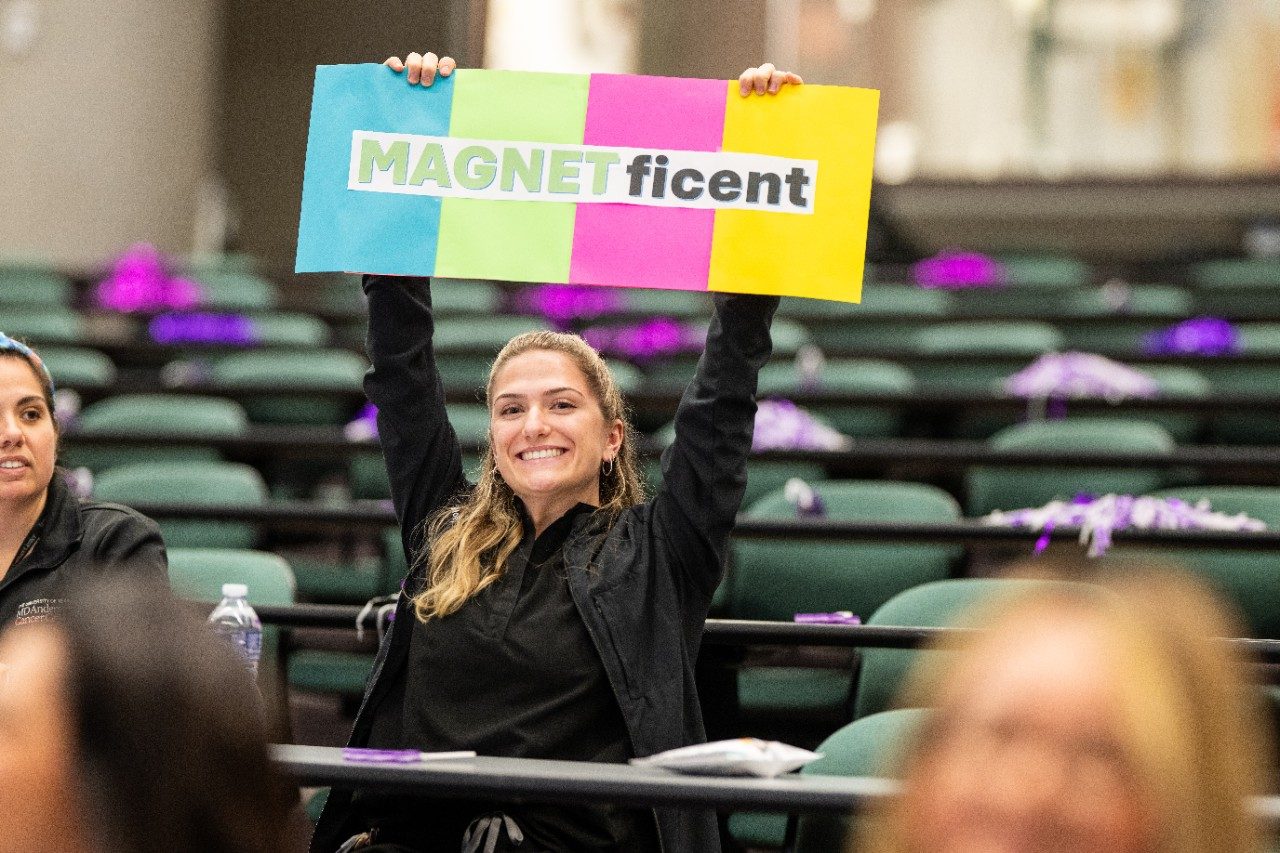 An MD Anderson nurse cheers and holds a multi-colored sign that reads magnet-ficent. 