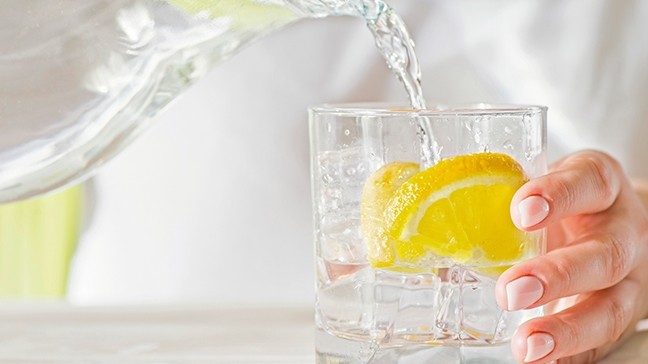 Person in white shirt pours water from a glass pitcher into a glass cup with ice and a lemon slice.