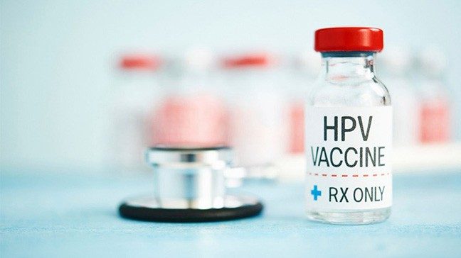 A glass vial with red cap and a label reading 'HPV VACCINE RX ONLY' on a blue surface with a stethoscope and additional vials in the background.