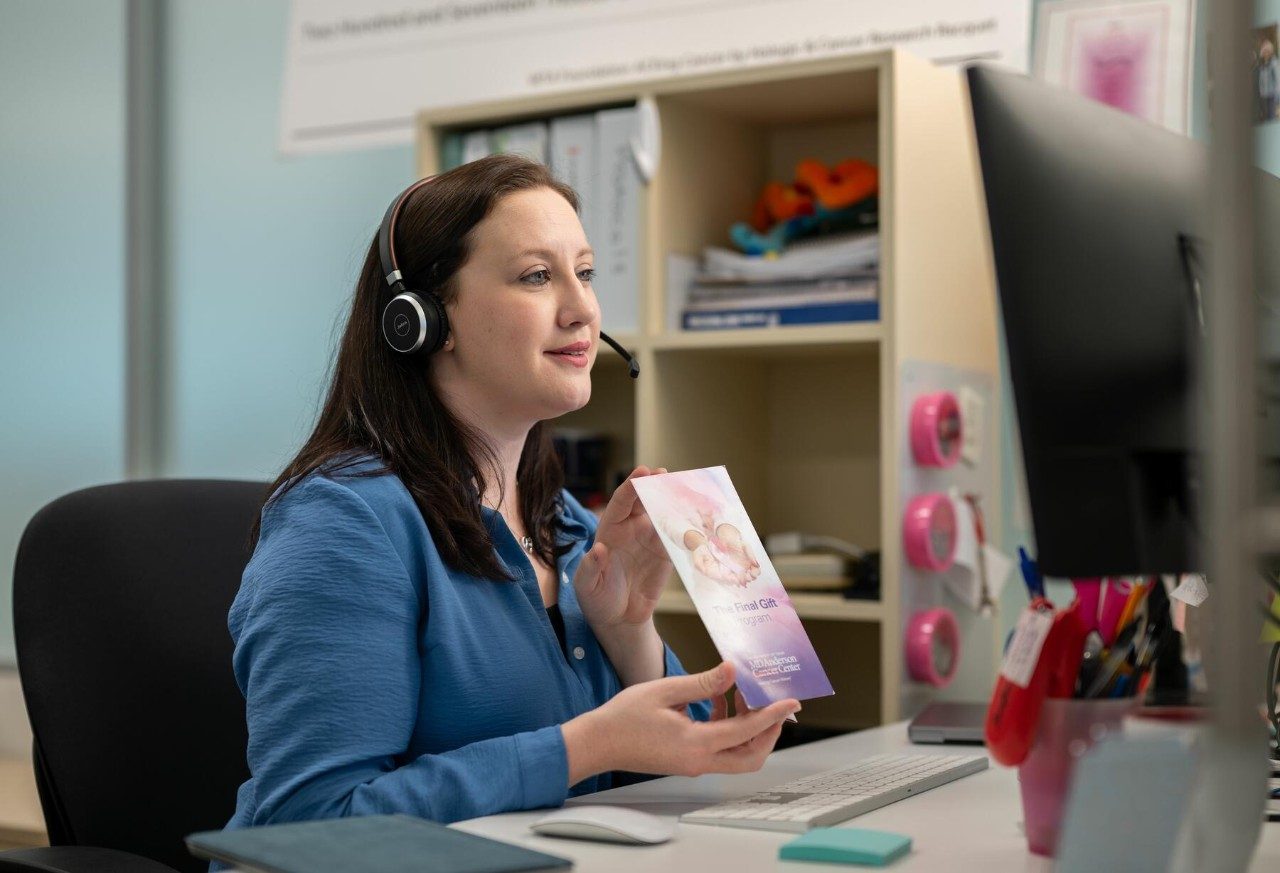 Woman in a blue shirt sits at a desk wearing headphones and holding a purple brochure.