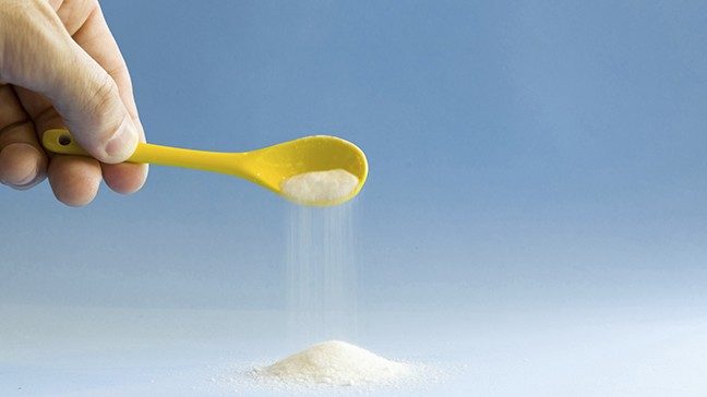Hand holds a yellow spoon pouring out white granulated sugar