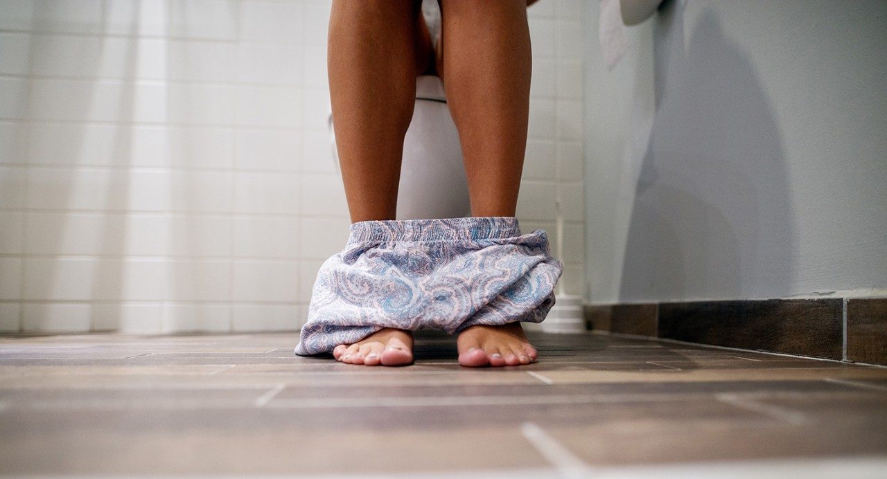 Photo shows person's feet and lower legs while sitting on a toilet.