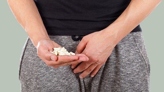 A man's right hand holds several white pills while the left hand rests on his groin area.