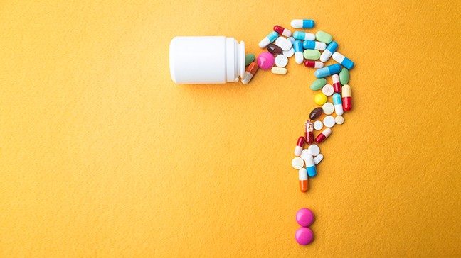 White pill bottle spills out a variety of colorful capsules and pills in the shape of a question mark against an orange background