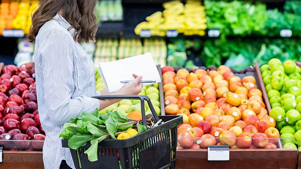 Woman shopping in produce section holding a black grocery basket and white grocery list.