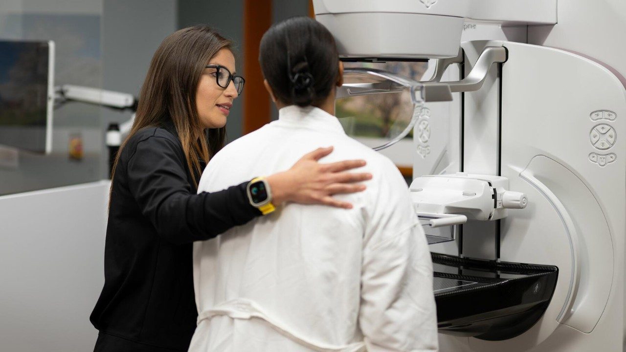 Woman places her hand on the back of a female patient wearing a white robe. Both are standing in front of a mammography machine.