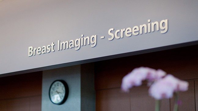 A grey wall with an analog clock and a sign below a sign reading 'Breast Imaging - Screening'